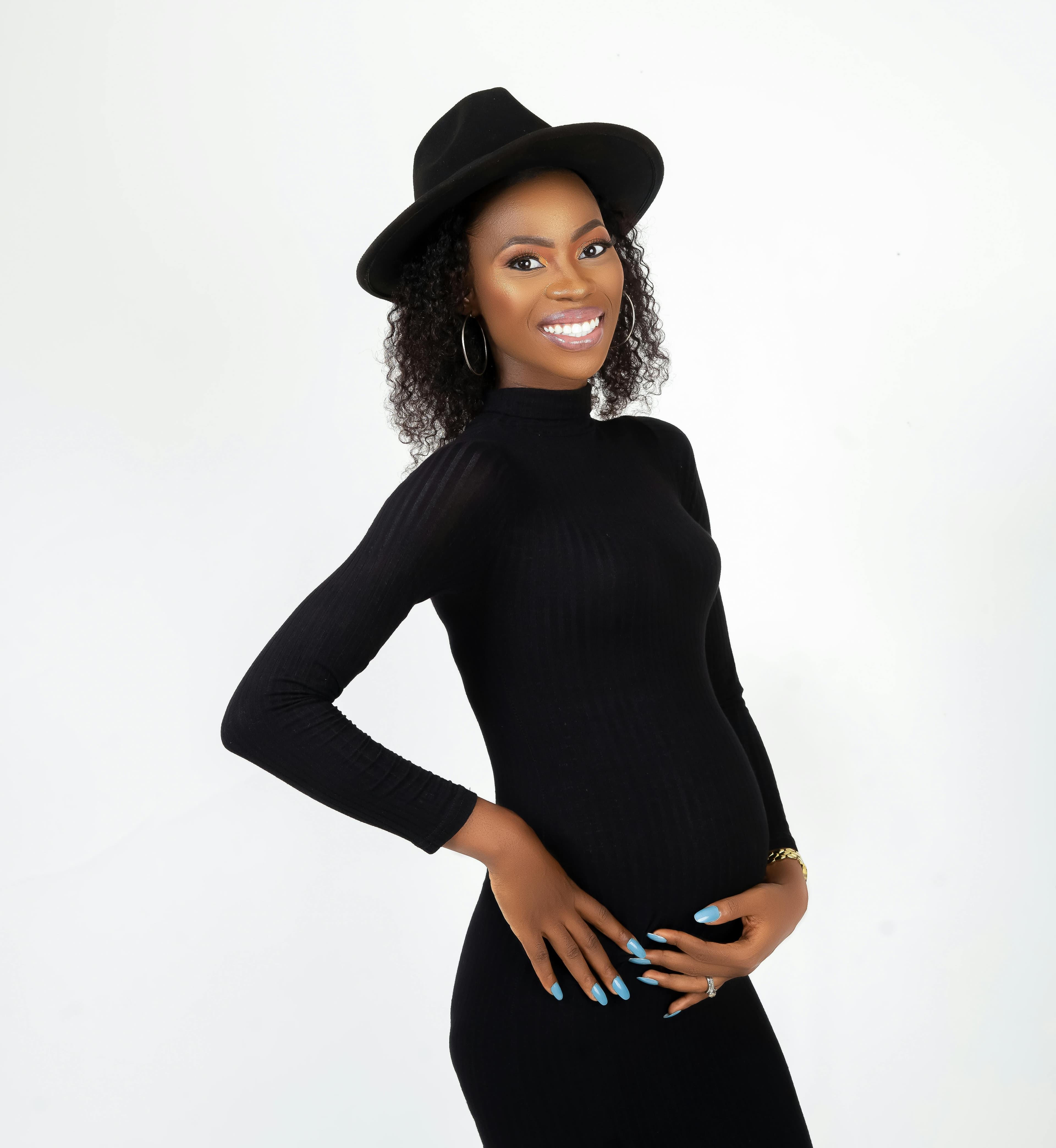 Femme enceinte souriante vêtue d'une robe noire ajustée et portant un chapeau noir, posant avec élégance devant un fond blanc.