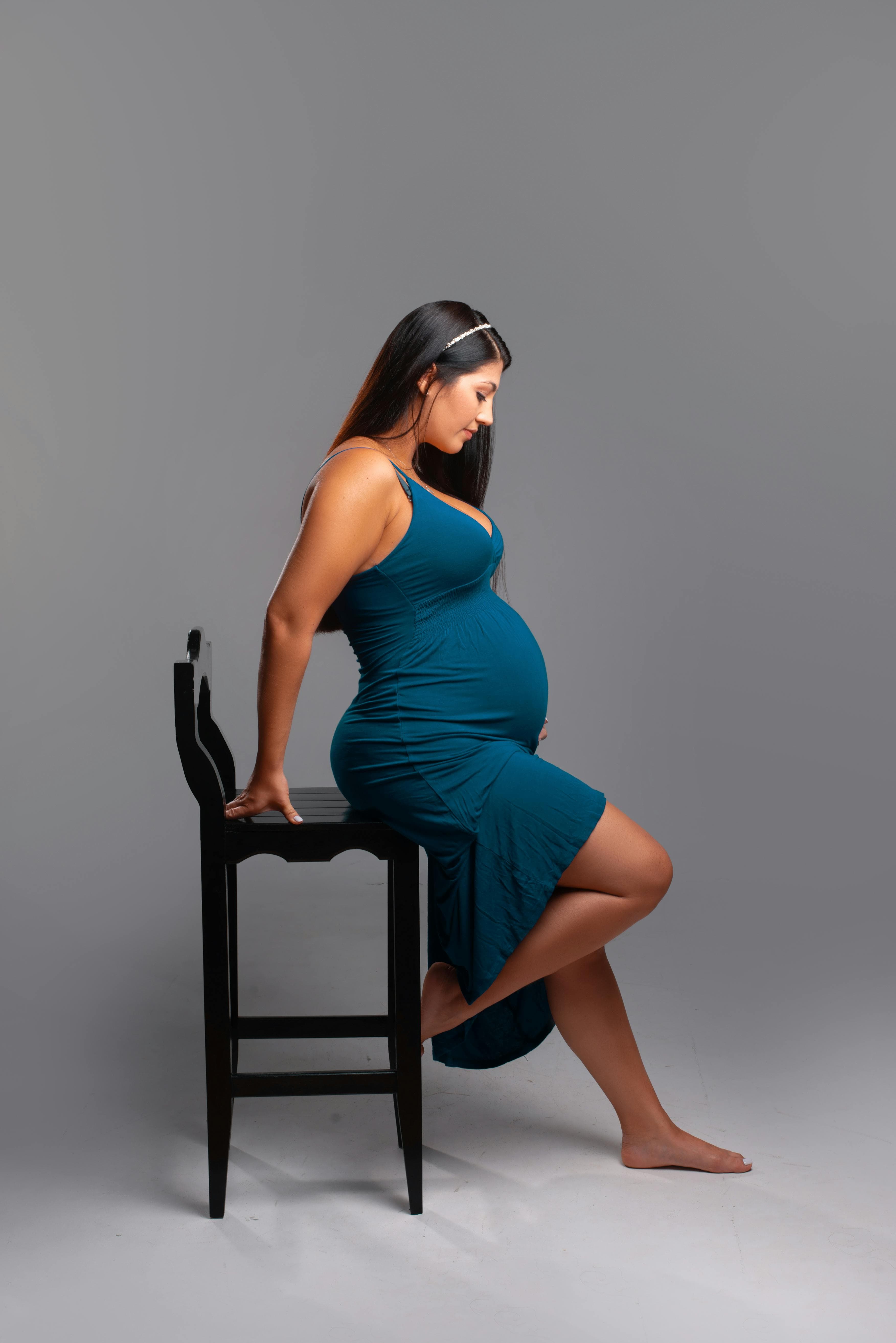 Femme enceinte portant une robe bleue ajustée, assise sur le bord d'une chaise noire dans un studio au fond gris neutre.