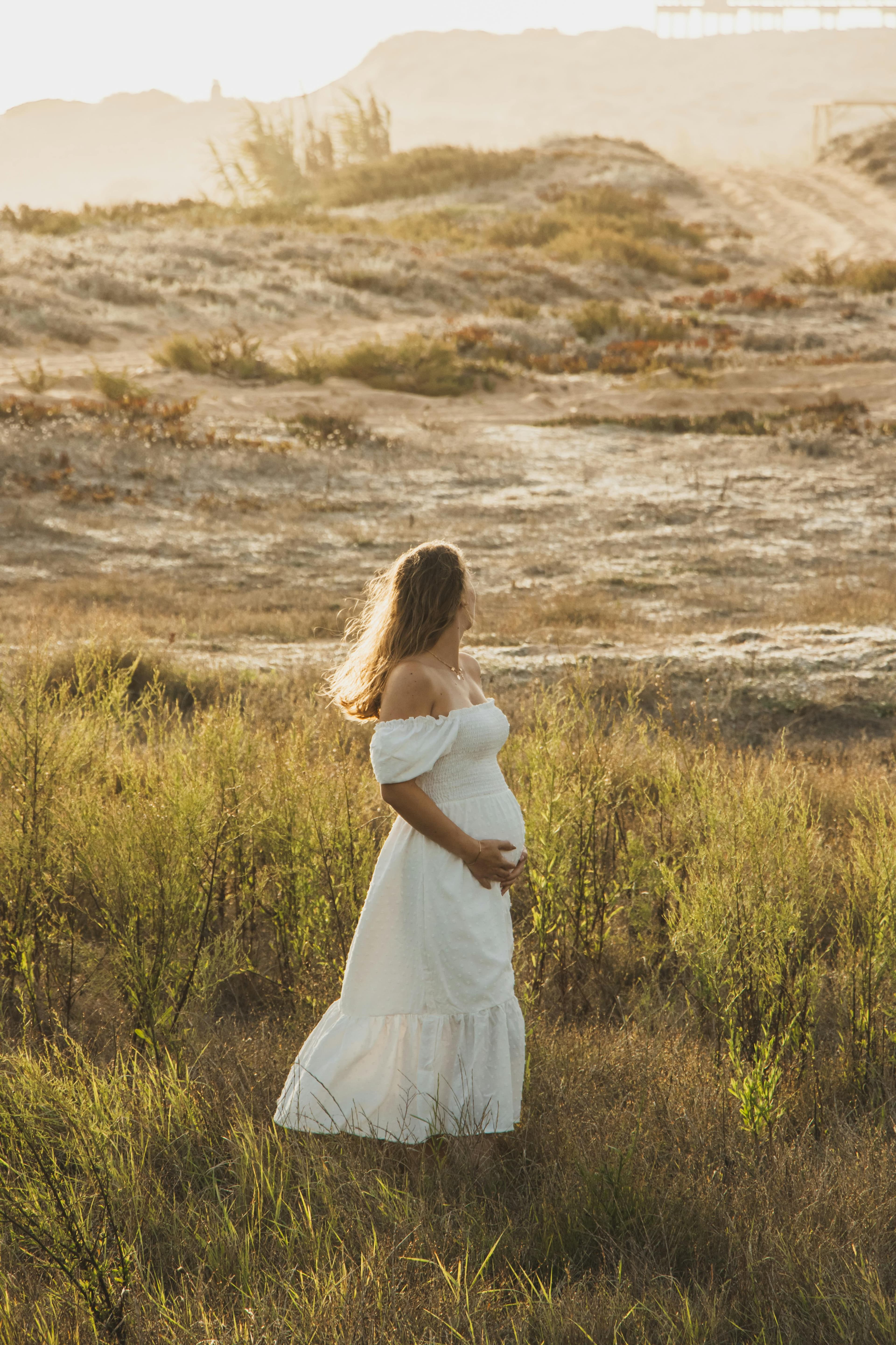 Femme enceinte dans une robe blanche, debout dans un champ ensoleillé, entourée de végétation naturelle et regardant vers l'horizon.