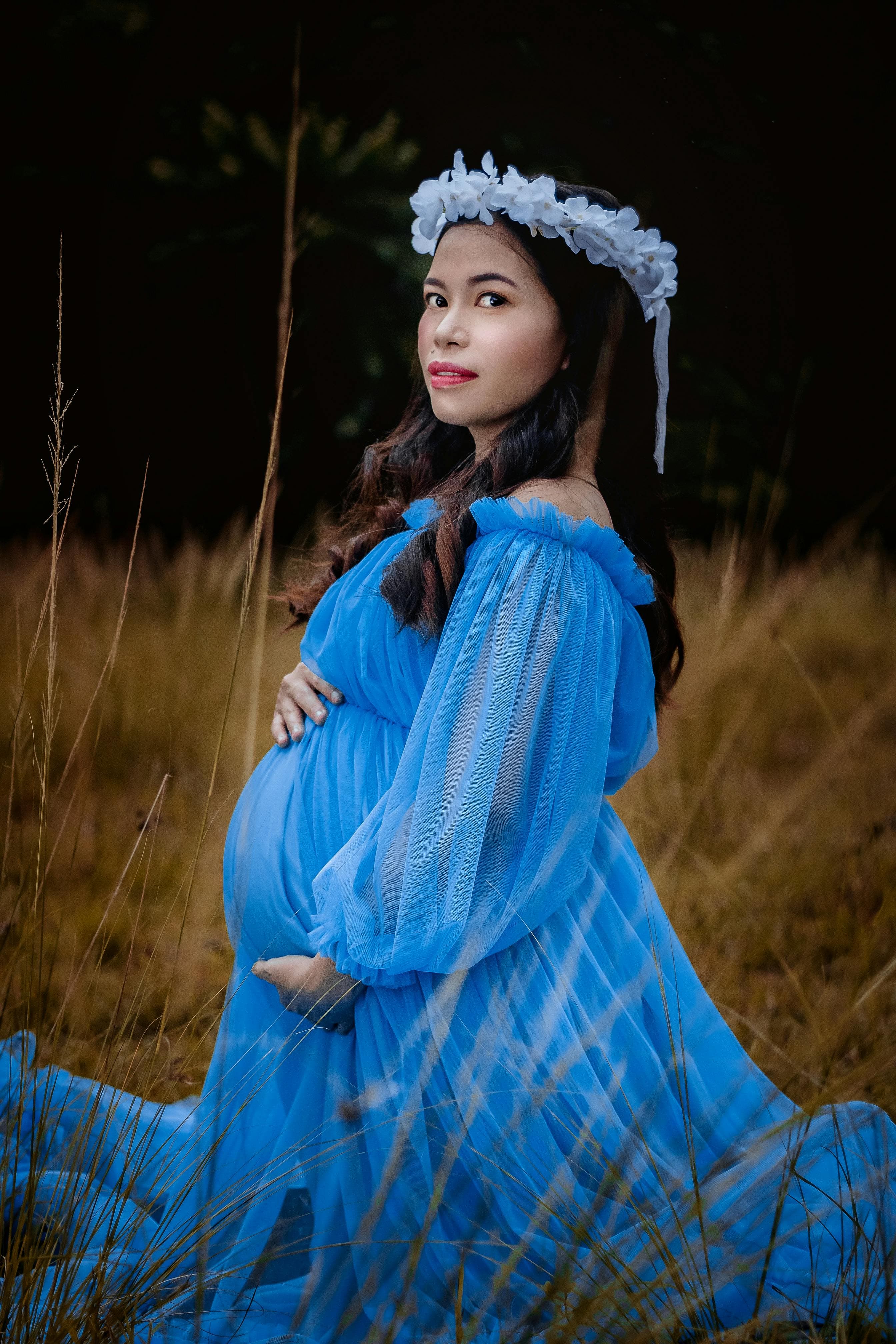 Femme enceinte portant une robe bleue fluide et une couronne de fleurs blanches, assise dans un champ herbeux avec un fond sombre.